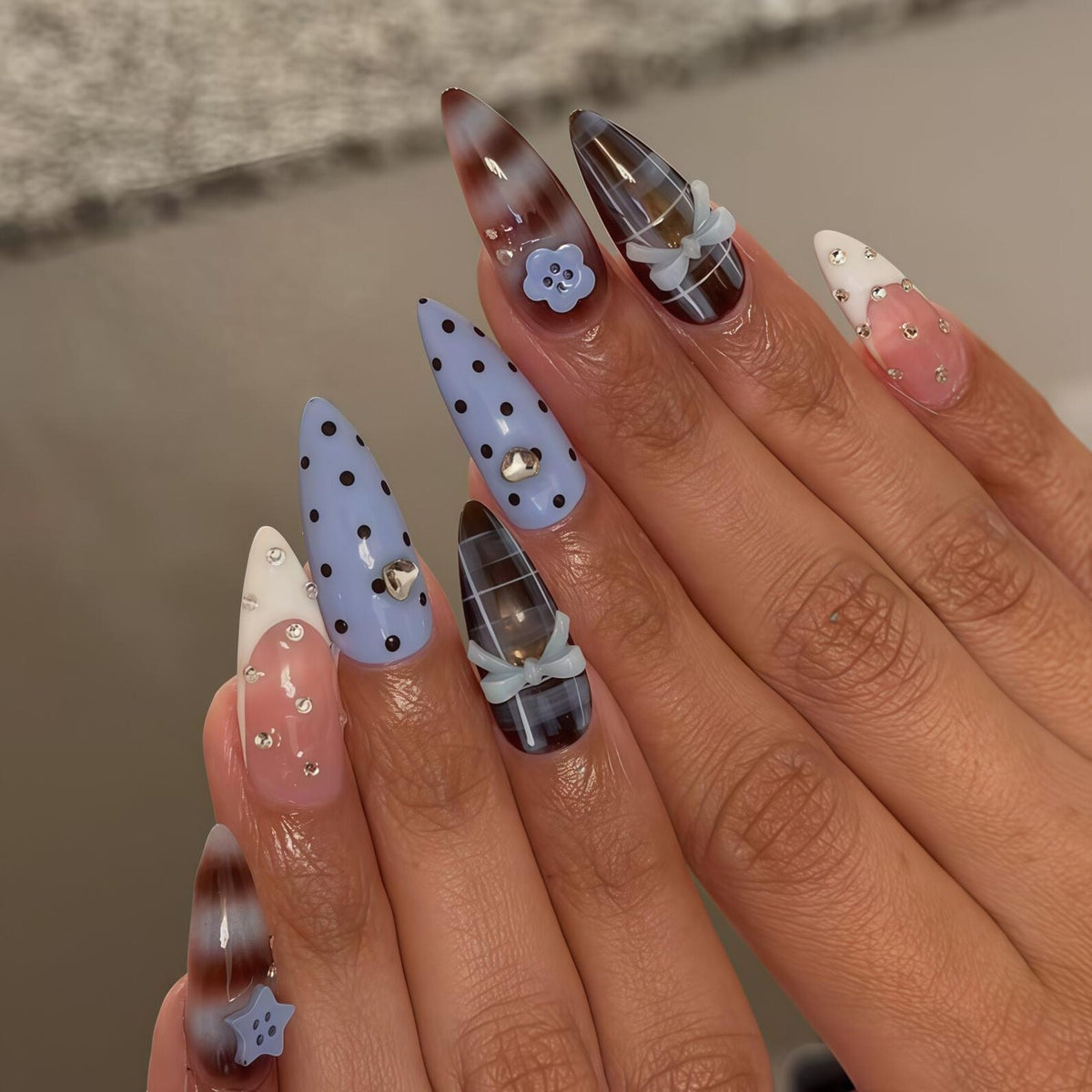 A set of long, almond-shaped acrylic nails. Designs include light blue polka dots, plaid, and a gradient brown and white. Embellishments include small bows, rhinestones, and a flower. The nails are a mix of colors and patterns.