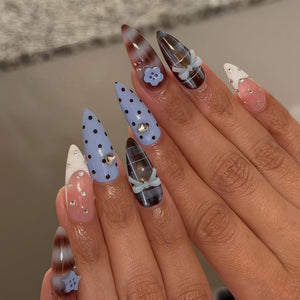 A set of long, almond-shaped acrylic nails. Designs include light blue polka dots, plaid, and a gradient brown and white. Embellishments include small bows, rhinestones, and a flower. The nails are a mix of colors and patterns.