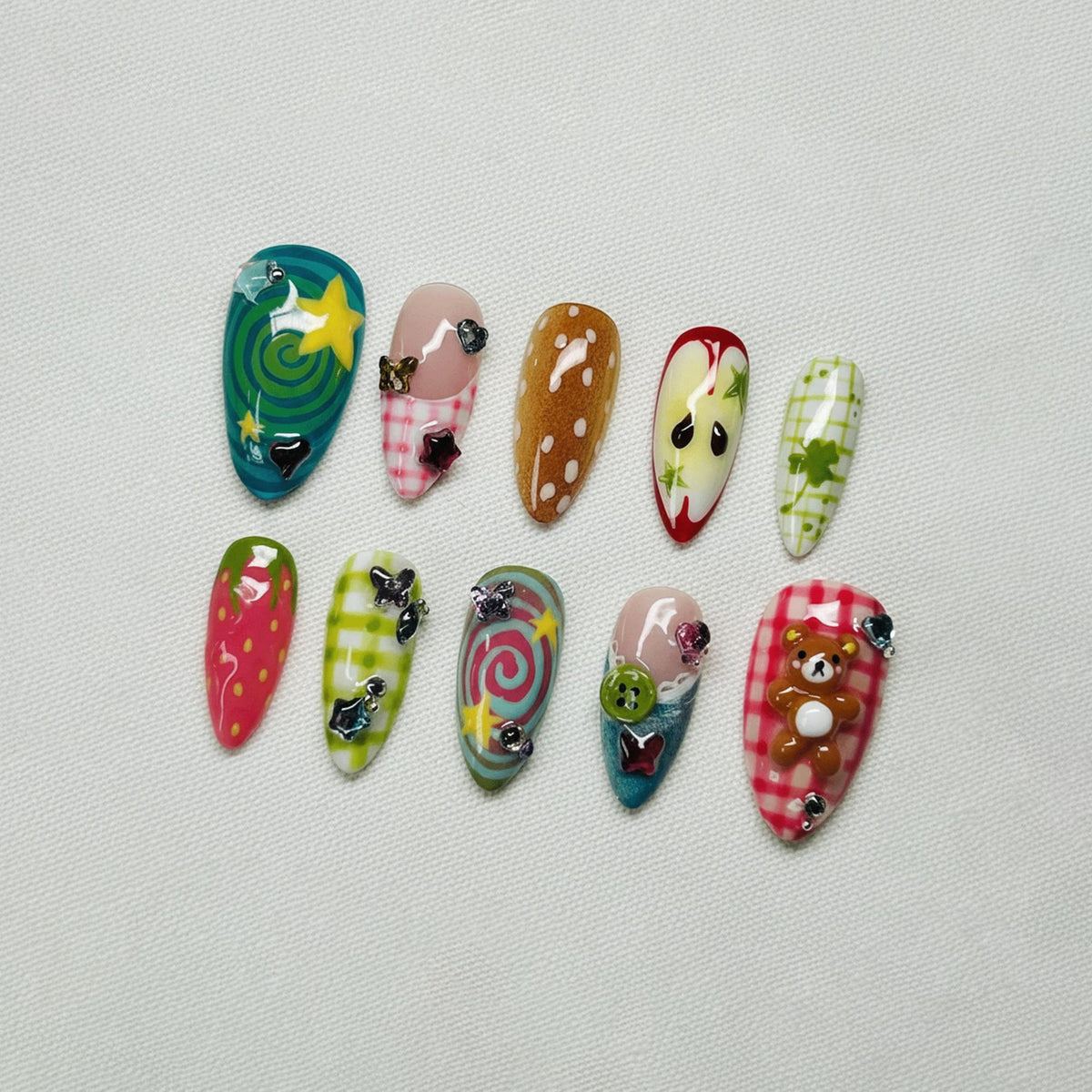 A collection of ten uniquely designed artificial nails. Each nail features a different design, including swirls, stars, polka dots, and a teddy bear. Colors include green, pink, red, and brown, with decorative embellishments.