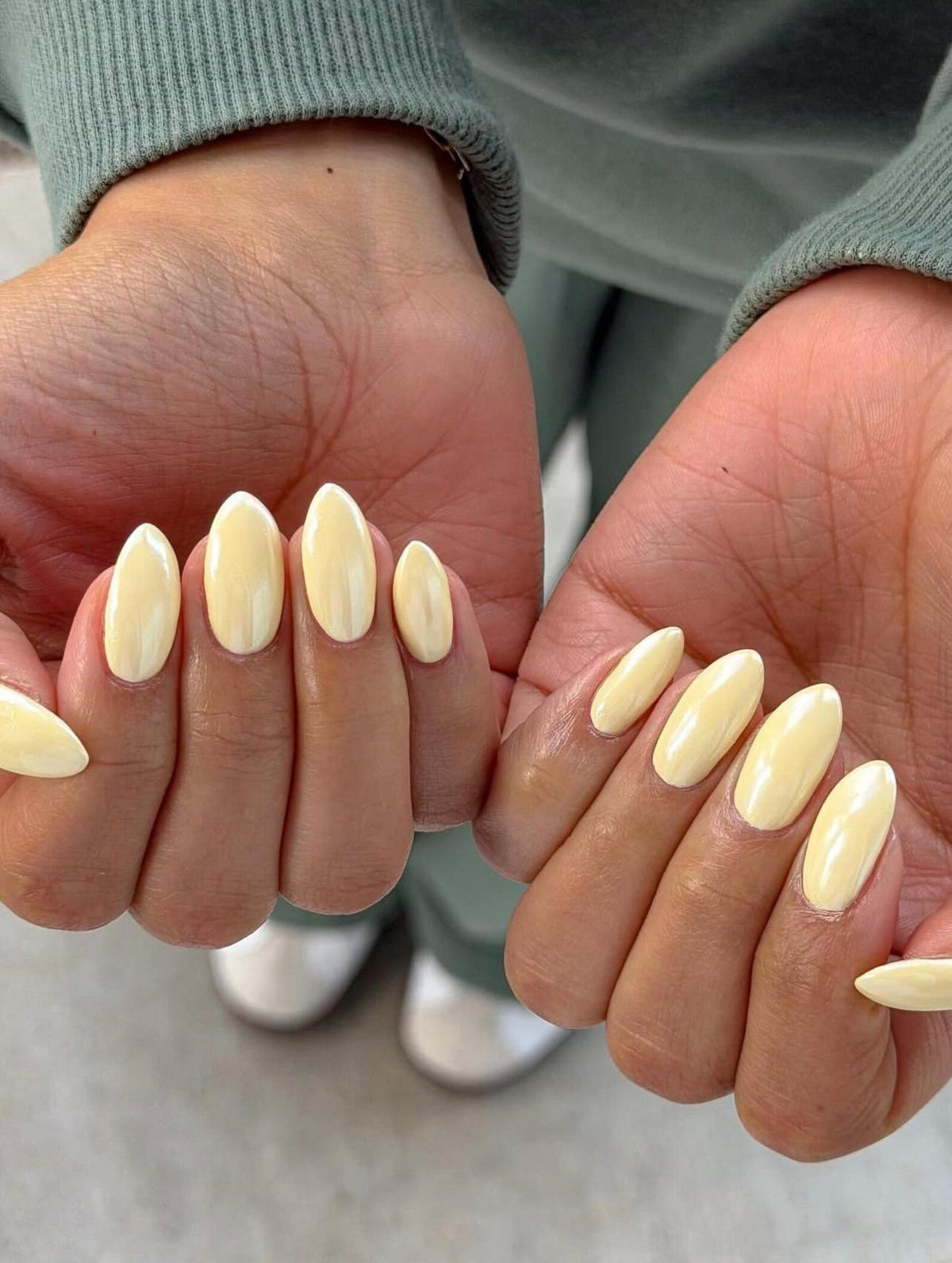 Close-up of two hands with oval-shaped, pale yellow nail polish. The nails are neatly manicured and have a glossy finish. The background is blurred, with hints of clothing and a light-colored surface.