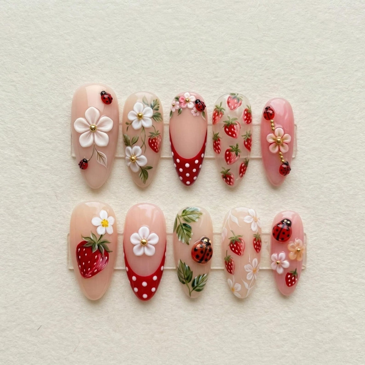 A set of ten press-on nails featuring a nude base with intricate 3D designs including red strawberries, white flowers, green leaves, and small ladybugs. Some nails display a red tip with white polka dots and gold accents.