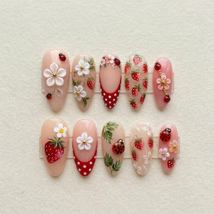 A set of ten press-on nails featuring a nude base with intricate 3D designs including red strawberries, white flowers, green leaves, and small ladybugs. Some nails display a red tip with white polka dots and gold accents.