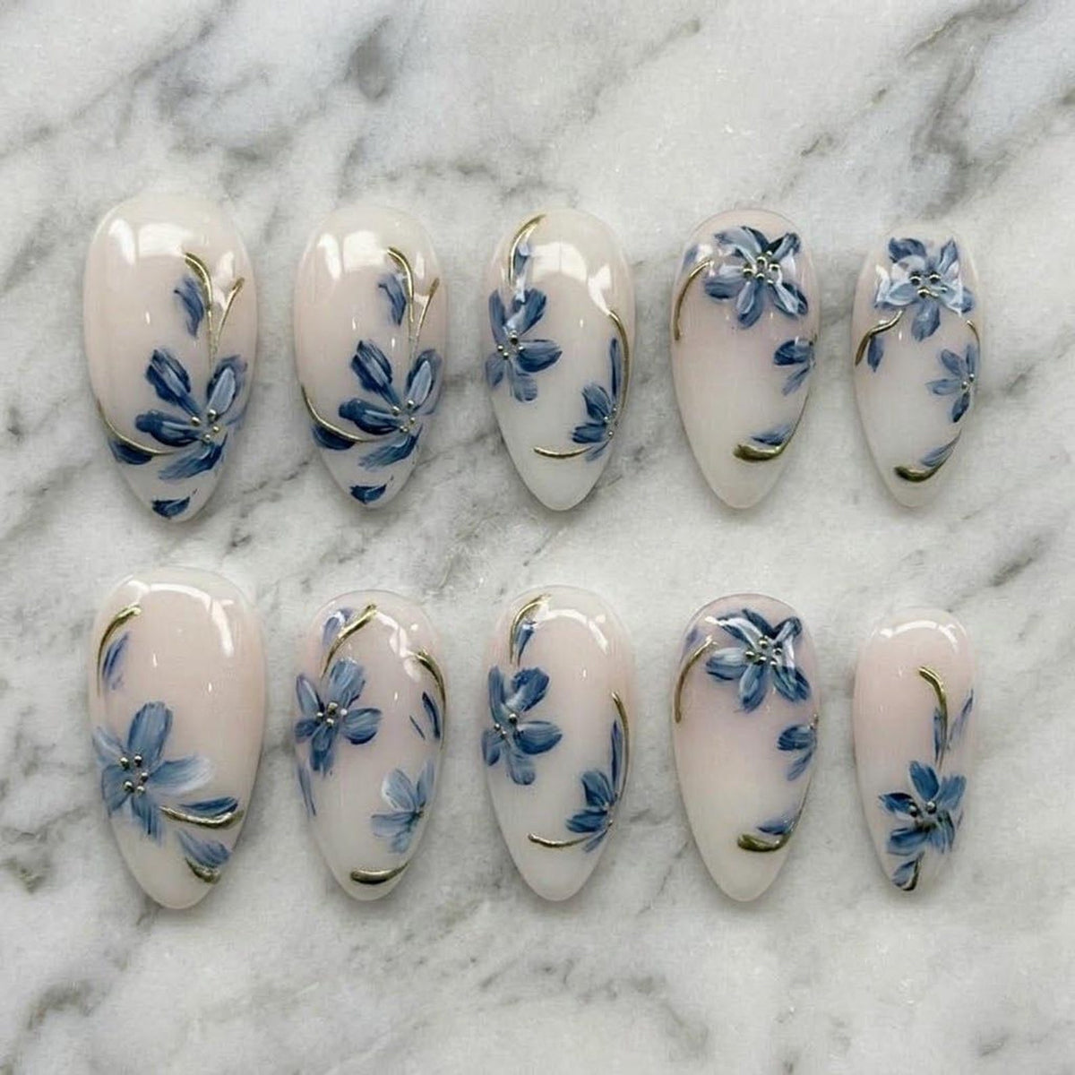 A set of ten oval-shaped artificial nails. Each nail is painted with a light pink base and features a blue floral design with gold accents. The nails are arranged on a white marble surface.