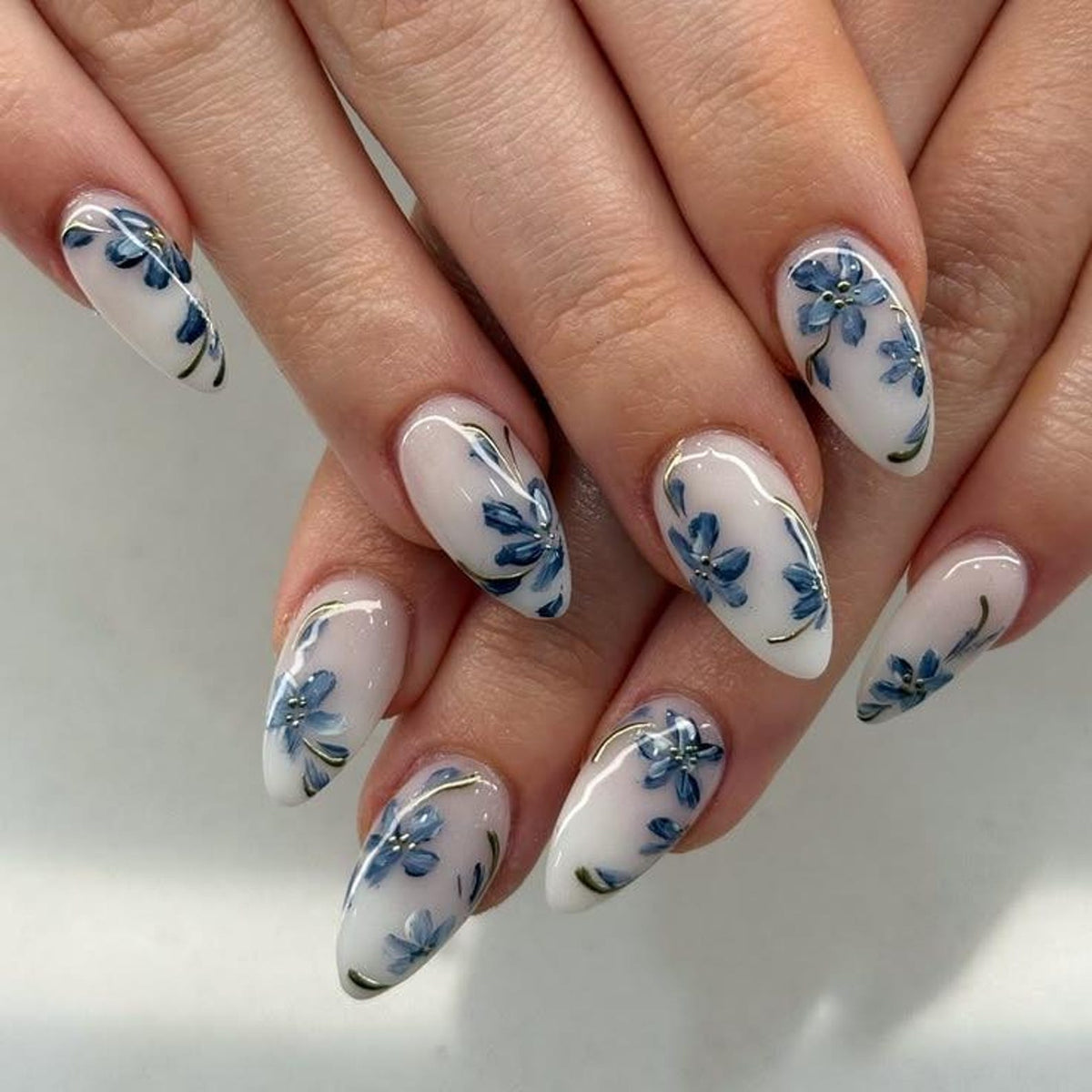 Almond-shaped nails with a white base and delicate blue floral designs. Each nail features a different arrangement of the blue flowers, accented with thin gold lines. The nails are well-manicured and polished.