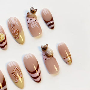 A collection of almond-shaped false nails. Some nails feature musical notes, while others have a cat figurine. The color palette includes shades of pink, brown, and beige, with some nails having a French tip design.