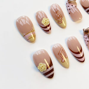 A set of almond-shaped false nails in various designs. Some nails feature a nude base with brown stripes, floral accents, or musical notes. Others have a nude base with a grid pattern or a brown outline.