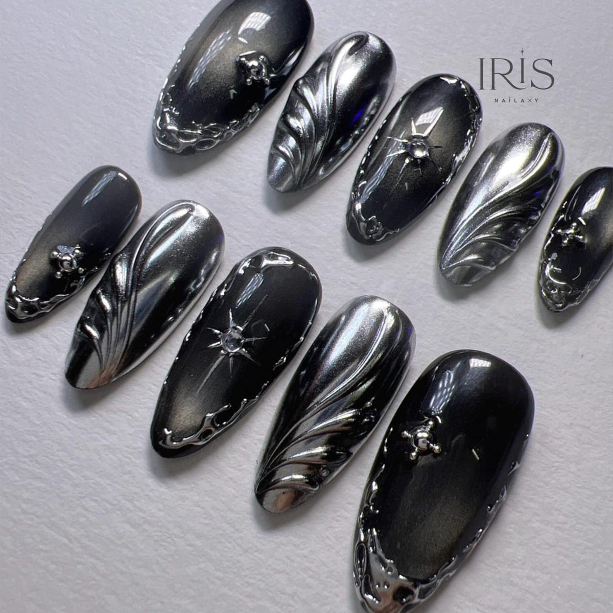 A set of ten long, oval-shaped artificial nails. The nails are painted in shades of black and silver with intricate silver designs, including swirls, starbursts, and decorative edges. The Iris Nailaxy logo is visible in the upper right corner.