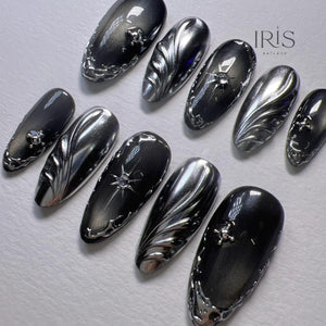 A set of ten long, oval-shaped artificial nails. The nails are painted in shades of black and silver with intricate silver designs, including swirls, starbursts, and decorative edges. The Iris Nailaxy logo is visible in the upper right corner.