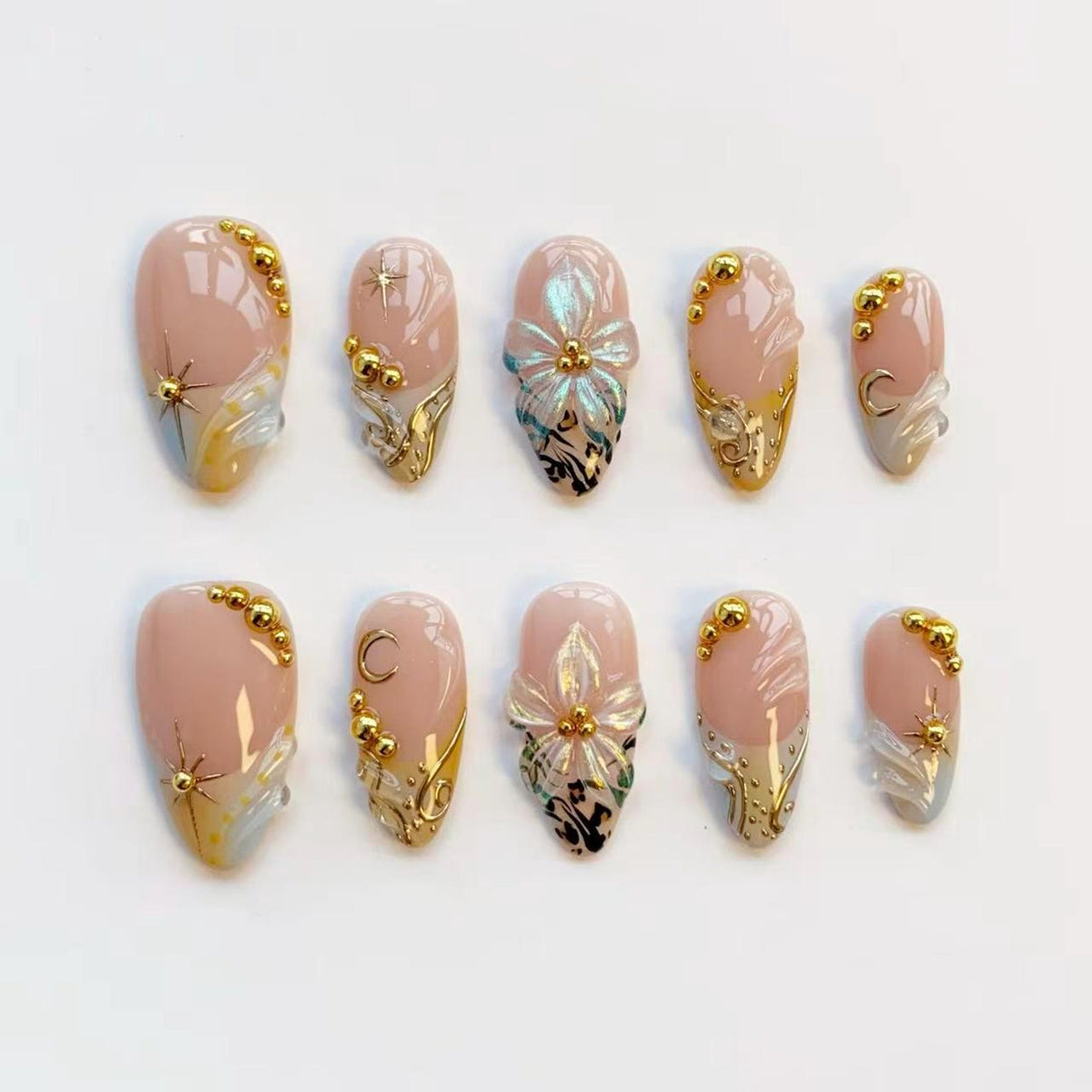 A set of ten artificial nails in various sizes. Each nail features a nude base with gold accents, including starbursts, crescent moons, and floral designs. Some nails have gold beads and intricate patterns.