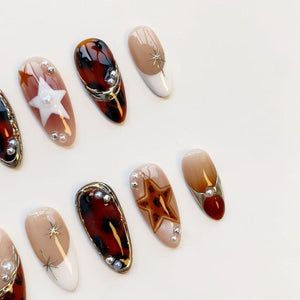 A collection of artificial nails with various designs. Some nails feature a tortoiseshell pattern, while others have star and pearl embellishments. The color palette includes shades of brown, beige, and white, with gold accents. The nails are oval-sh
