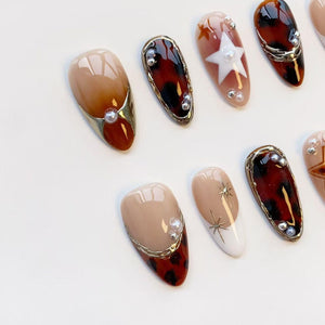A collection of almond-shaped artificial nails in various designs. Some nails feature a tortoiseshell pattern, gold trim, and pearl accents. Others have a nude base with star or geometric designs. The color palette includes brown, beige, and white.