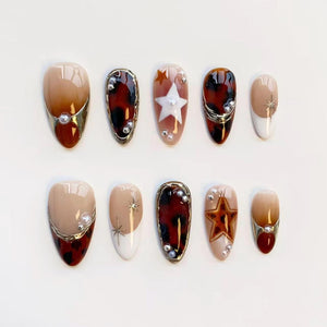 A set of ten almond-shaped press-on nails. Designs include tortoiseshell, gold accents, pearls, and star embellishments. Colors include beige, brown, white, and gold. The nails are arranged on a white surface.