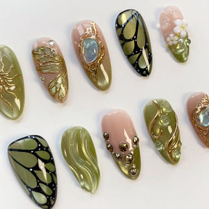A collection of professionally designed artificial nails. The nails feature various designs, including gold embellishments, butterfly patterns, and floral accents. Colors include olive green, pink, and gold, with a glossy finish. The nails are arrang