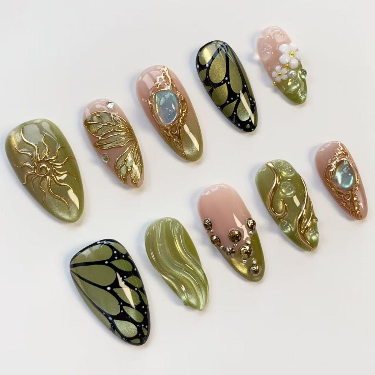 A collection of ten artificial nails with various designs. The nails feature a range of colors, including olive green, pink, and gold. Some nails have intricate gold embellishments, while others showcase butterfly wing patterns and gem accents.
