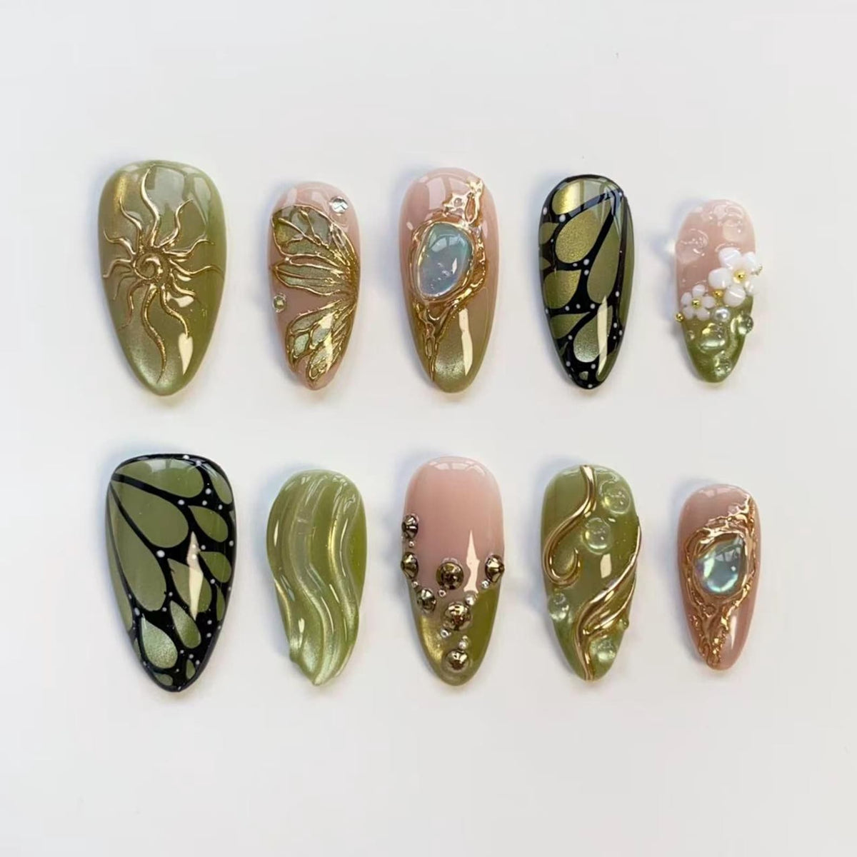 A collection of ten artificial nails in various designs. The nails feature gold accents, including sunbursts, butterfly wings, and floral patterns. Colors include olive green, pink, and gold, with some nails showcasing a glossy finish.