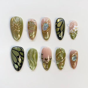 A collection of ten artificial nails in various designs. The nails feature gold accents, including sunbursts, butterfly wings, and floral patterns. Colors include olive green, pink, and gold, with some nails showcasing a glossy finish.