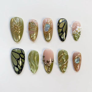 A collection of ten artificial nails in various designs. The nails feature a range of colors, including olive green, pink, and gold. Some nails have intricate gold embellishments, while others showcase butterfly and floral patterns.