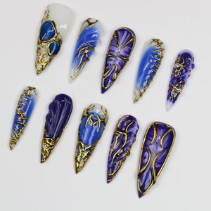 A collection of ten long, pointed artificial nails. Each nail features a unique design with shades of blue, white, and gold. The designs include swirls, embedded stones, and intricate gold filigree, creating an elegant and artistic aesthetic.