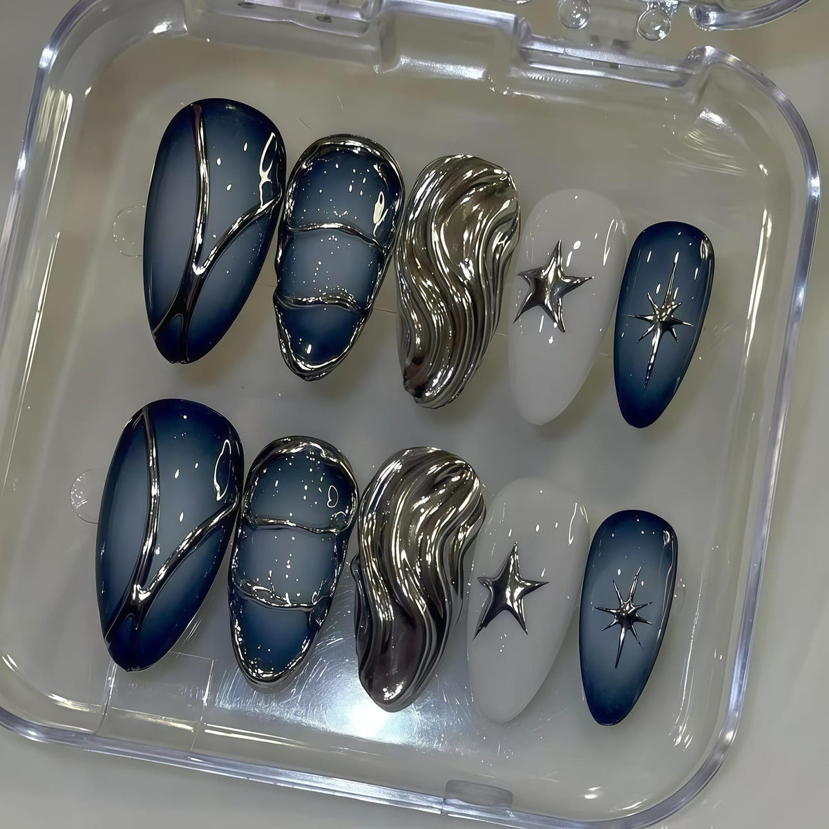 A set of ten artificial nails in a clear plastic case. The nails feature a variety of designs, including a gradient blue and silver swirl, silver wave, white with a star, and a dark blue with a starburst.