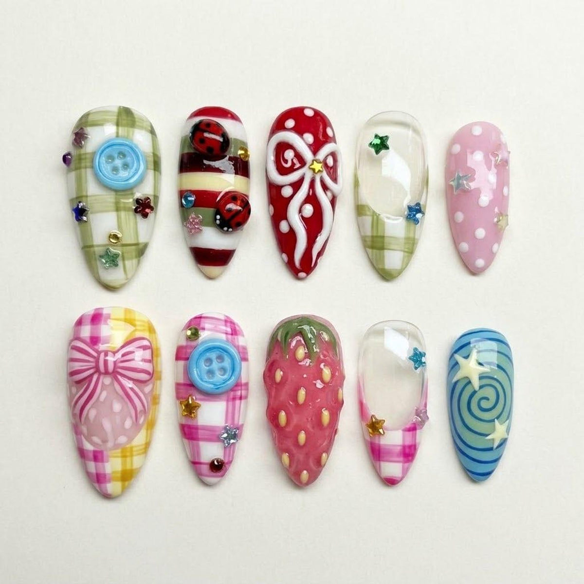 A collection of ten artificial nails with various designs. The nails feature patterns like gingham, polka dots, and bows, along with embellishments such as buttons, ladybugs, and stars. Colors include red, pink, blue, green, and yellow.