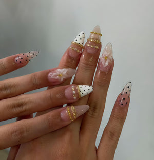 A set of long, pointed acrylic nails. Some nails feature a clear base with black polka dots, while others have a white base with black polka dots. Gold rings and floral accents adorn some nails, adding a touch of elegance.