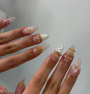 Close-up of manicured nails with a variety of designs. Some nails are clear with white polka dots, others are adorned with gold rings, pearls, and floral accents. The nails are long and pointed, showcasing intricate nail art.