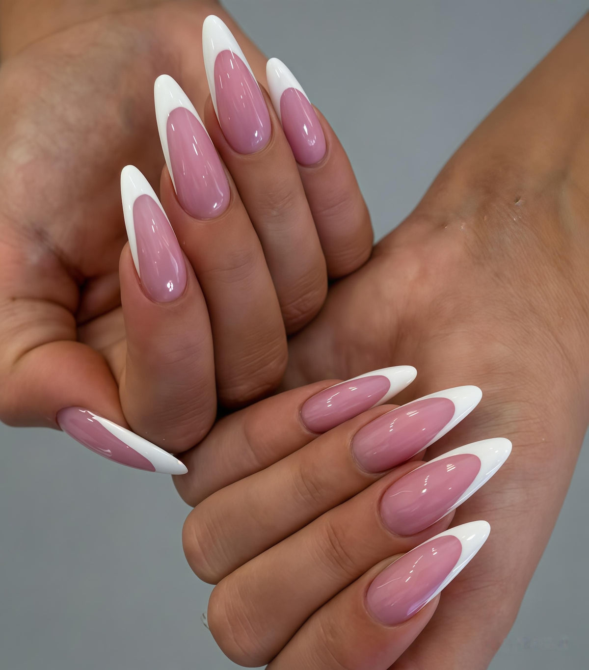 A close-up of two hands displaying long, almond-shaped nails. Each nail features a white tip and a soft pink base, creating a classic French manicure style. The nails are glossy and well-manicured.