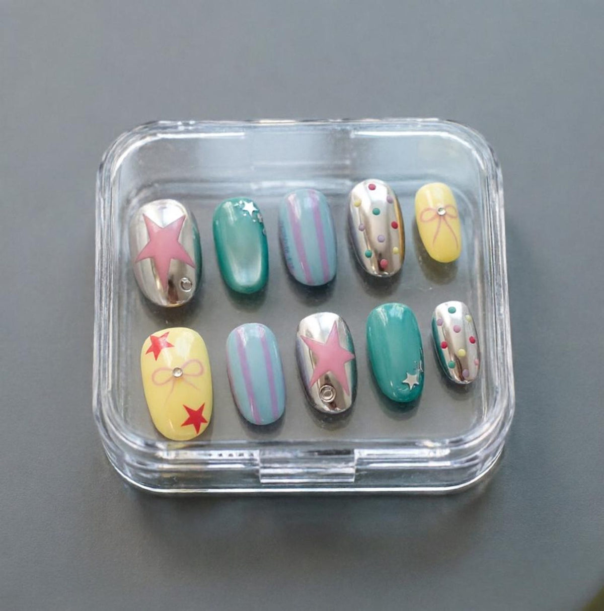 A set of ten artificial nails in a clear plastic case. The nails feature various designs, including silver metallic bases with pink stars, teal, and yellow with bows and red stars. Some nails have stripes or small colorful dots.