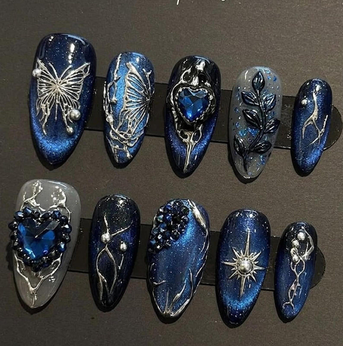 A set of ten artificial nails in various designs. The nails feature a dark blue glitter base with silver embellishments, including butterflies, hearts, and floral patterns. Some nails have a gray base with blue accents.