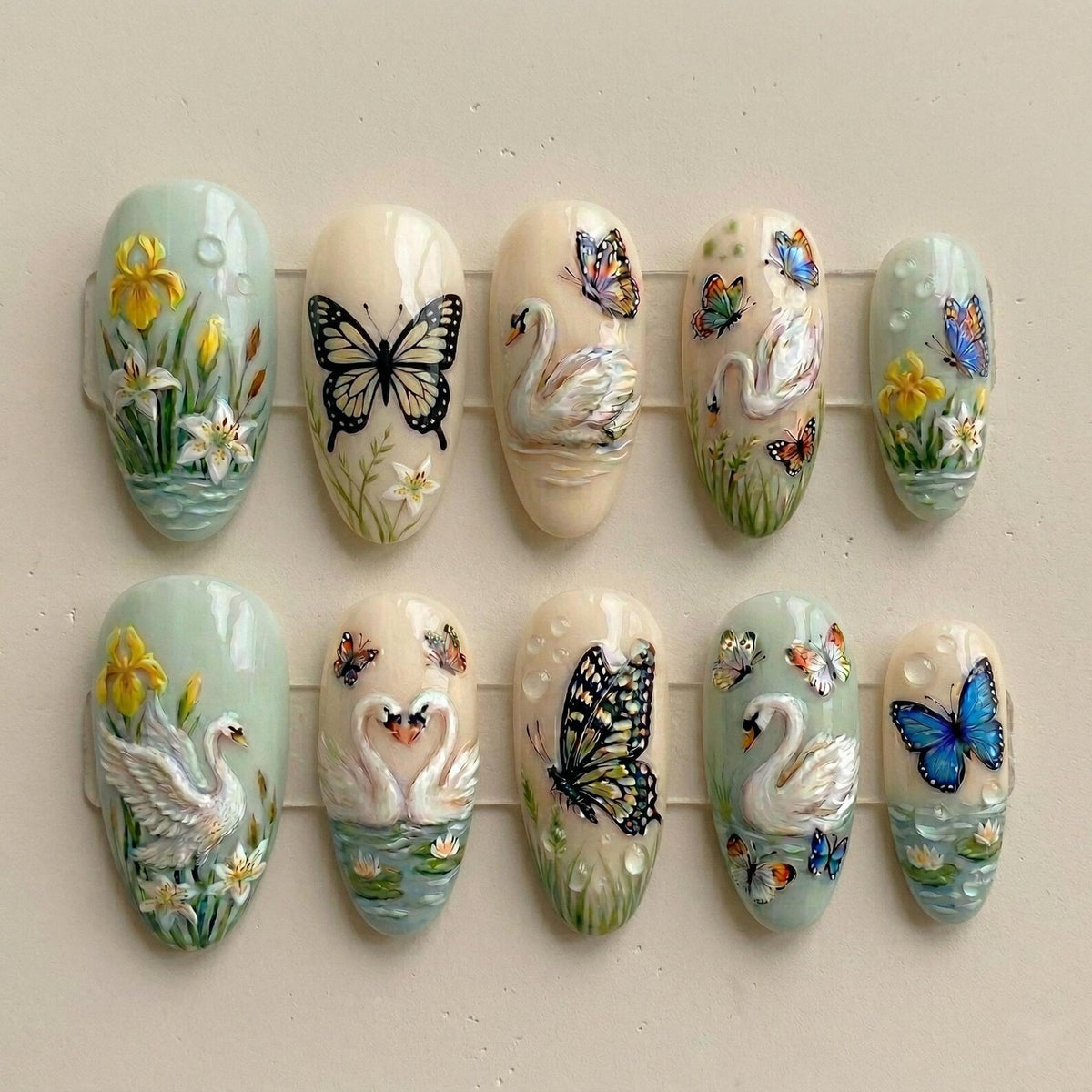 A set of ten artificial nails with detailed, hand-painted designs. The nails feature butterflies, swans, and floral elements in a variety of colors, including blue, yellow, and white. The nail shapes are oval.