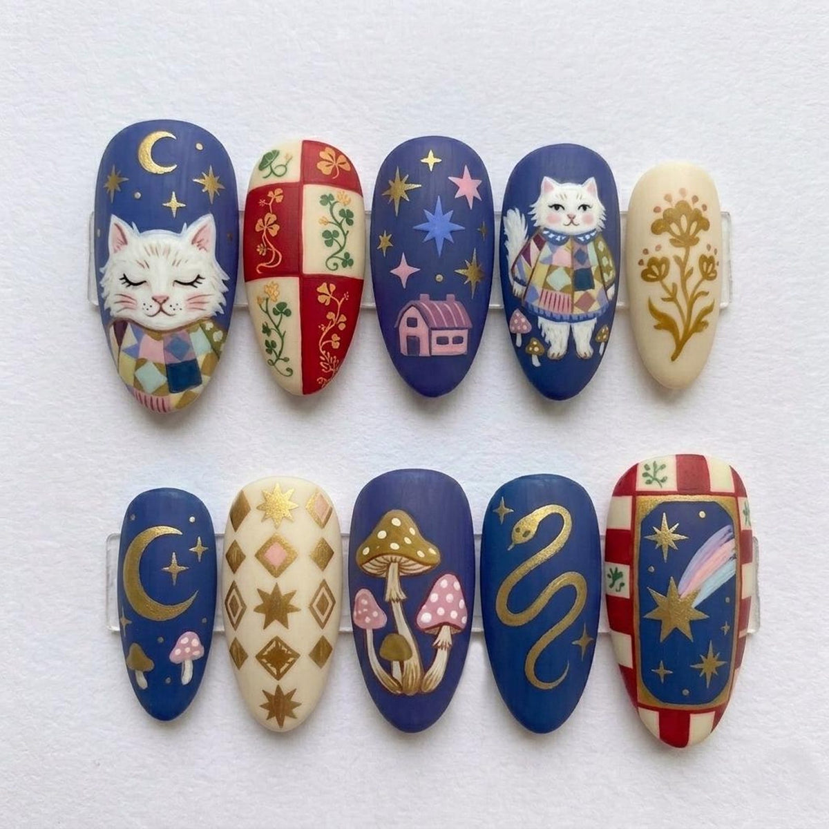 A set of ten almond-shaped false nails with whimsical designs. The nails feature illustrations of a cat, mushrooms, stars, a house, a snake, and floral patterns in a palette of blue, gold, red, and cream.