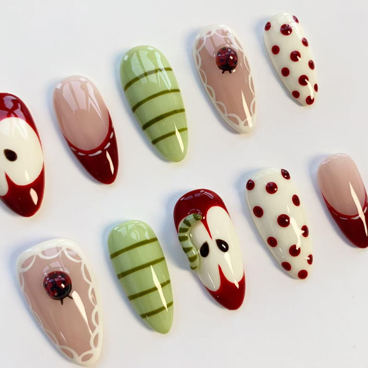 A collection of artificial nails with various designs. Some nails feature red and white patterns, including polka dots and apple-like shapes. Others have green stripes or a ladybug design on a pink base.