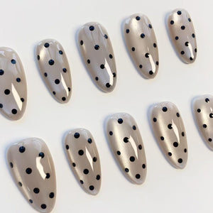 A set of ten almond-shaped false nails. Each nail is painted a light beige color and decorated with evenly spaced black polka dots. The nails have a glossy finish and are arranged on a white surface.