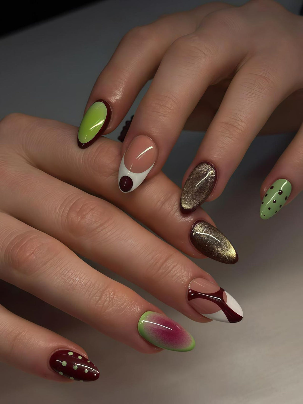 A close-up of a hand with almond-shaped nails. The nails feature various designs, including green with dark red trim, gold glitter, and a watermelon-inspired design. Other nails have white and dark red accents.