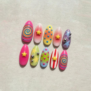 A set of ten colorful, almond-shaped artificial nails. Each nail features unique designs, including swirls, stars, polka dots, and stripes, in shades of pink, yellow, blue, and purple. Some nails have small, silver embellishments.