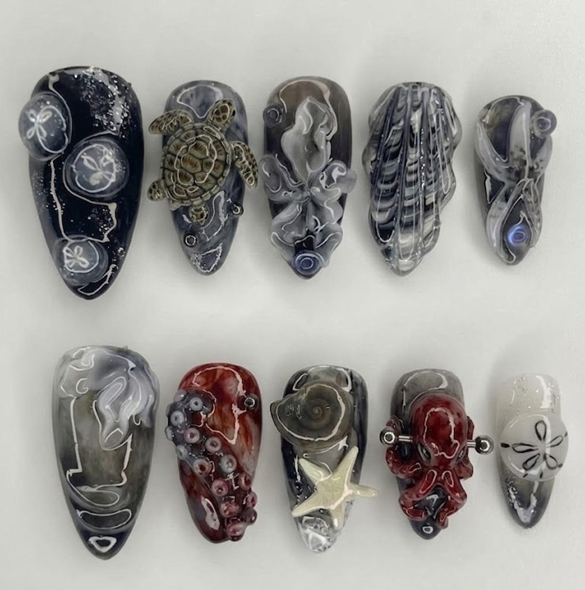 A set of ten artificial nails with various ocean-themed designs. The nails feature sea creatures like a turtle, octopus, and starfish, along with sand dollars and shell patterns. The color palette includes shades of gray, red, and white.