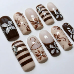 A set of artificial nails with various designs. Some nails are solid dark brown, others are beige with brown floral patterns or stripes. Several nails feature small, sparkling embellishments.