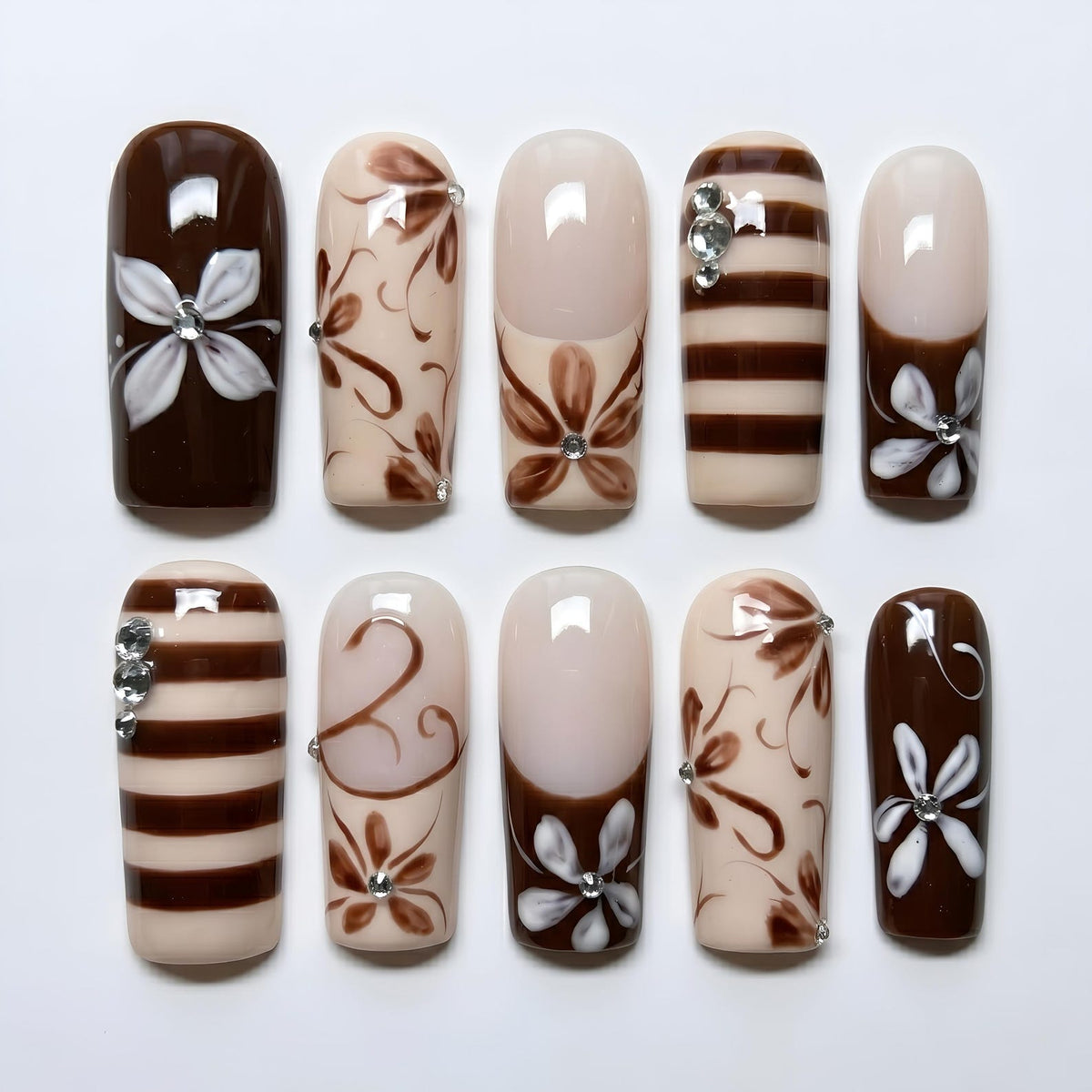 A set of ten artificial nails with various designs. The nails feature a mix of brown, beige, and white colors. Some nails have floral patterns, stripes, or a French tip design. Rhinestones are used as accents on some nails.