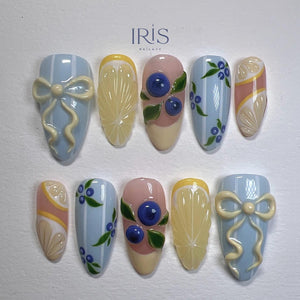 A set of ten almond-shaped artificial nails with various designs. The designs include light blue nails with white bows, lemon slices, blueberries with green leaves, and pink and yellow accents. The brand name "IRIS NAILAVY" is at the top.