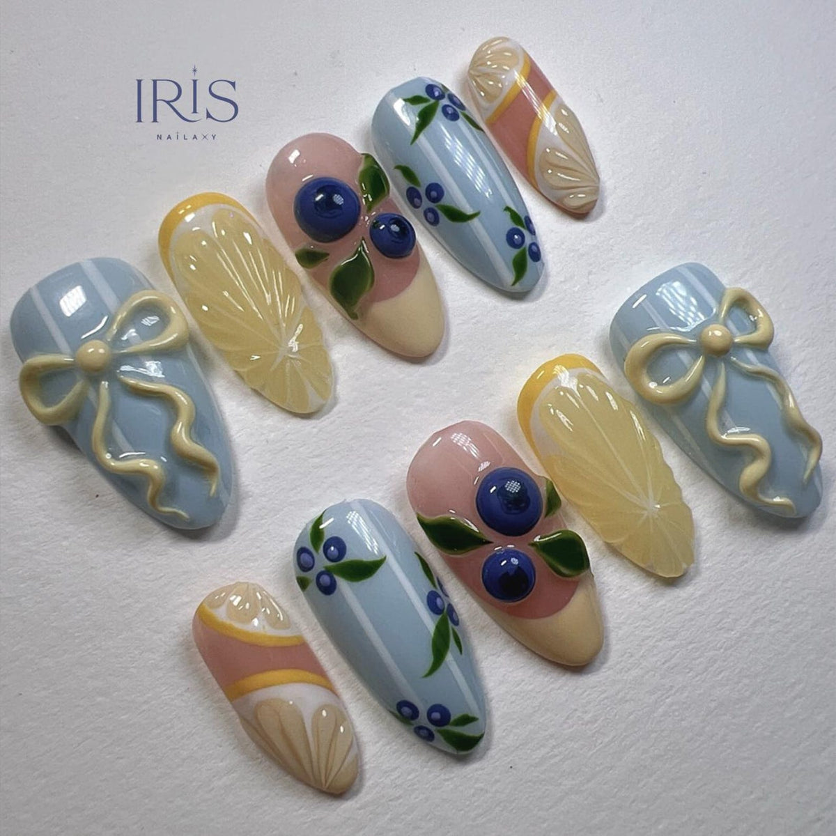 A collection of almond-shaped artificial nails with various designs. Some nails feature light blue bows, while others showcase lemon slices, blueberries, and striped patterns. The color palette includes light blue, yellow, pink, and white.