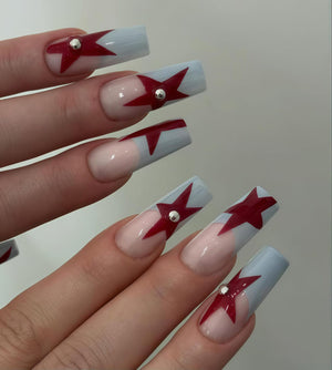 Long, square-shaped fingernails with a light blue and clear base. Each nail features a red star design and a small silver stud. The nails are well-manicured and have a glossy finish.