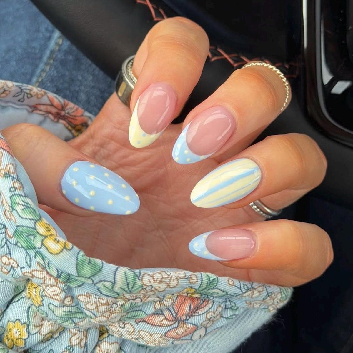 Almond-shaped nails featuring a mix of designs including light blue with white polka dots, pale yellow French tips with white dots, and pale yellow and light blue vertical stripes, all set against a natural nail base with silver rings.