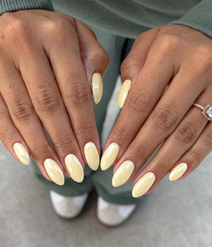 Close-up of hands with oval-shaped, pale yellow nail polish. A diamond ring is visible on one hand. The nails have a glossy finish. The background shows a person wearing a green sweater and pants.