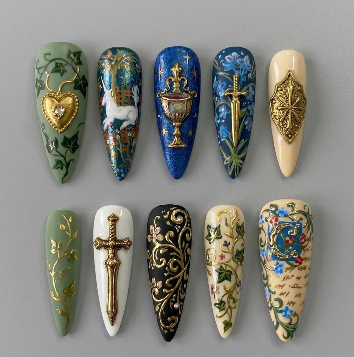A set of ten long, almond-shaped press-on nails with intricate, hand-painted designs. The nails feature a variety of colors, including green, blue, black, and cream, with gold accents. Designs include hearts, unicorns, swords, and floral patterns.
