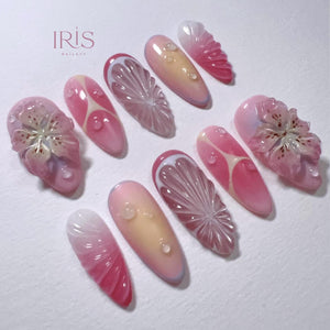A collection of artificial nails with various designs. Some nails feature a gradient of pink and white, while others showcase a carved citrus fruit design. Several nails have a 3D floral design, and all have water droplet accents. The brand name "IRI