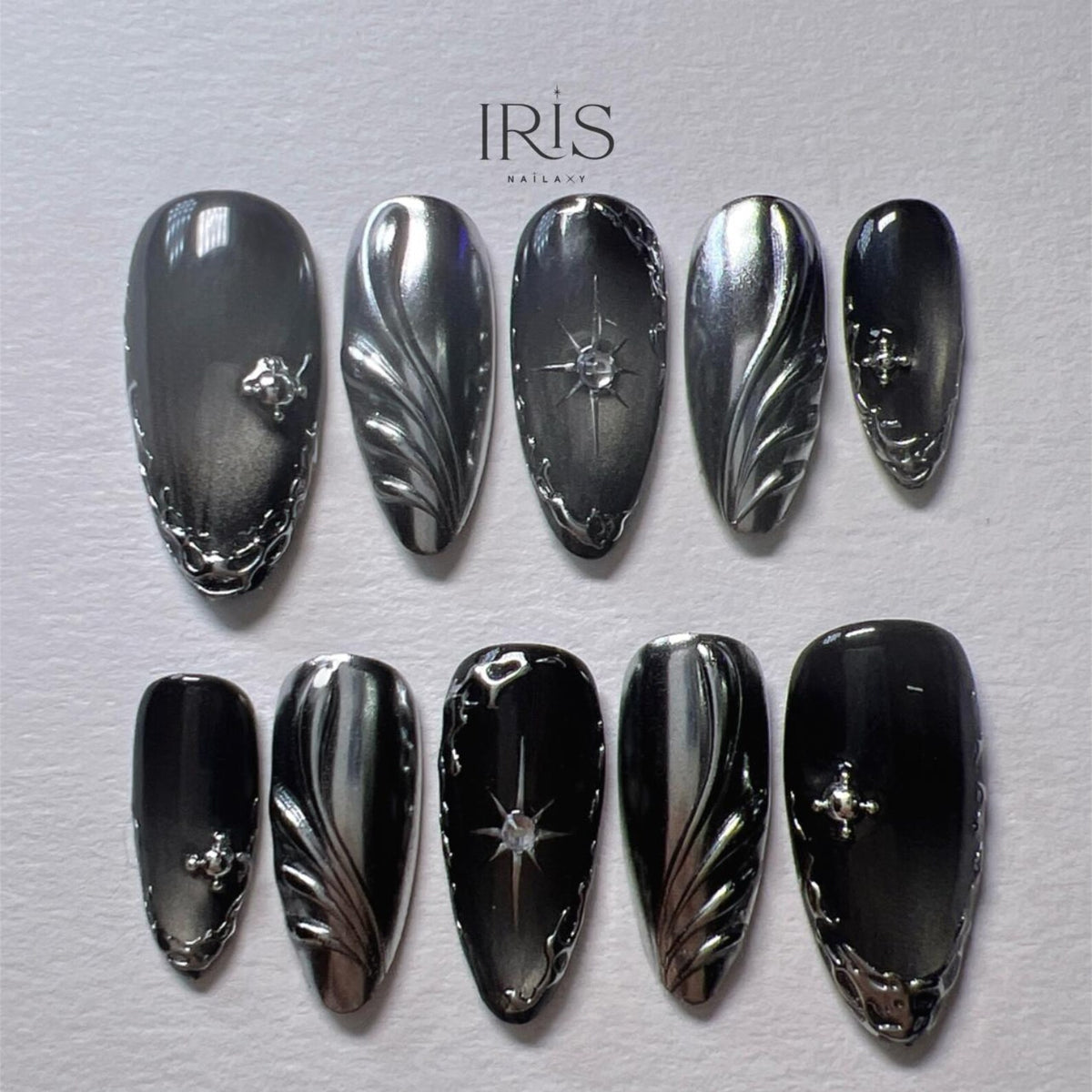 A set of ten press-on nails in a dark gray and silver color scheme. The nails feature intricate silver designs, including floral patterns, starbursts, and decorative edges. The brand name "IRIS NAILART" is visible at the top.