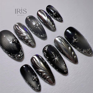 A set of ten long, oval-shaped artificial nails. The nails are a mix of silver and black, with intricate metallic designs. Some nails feature a starburst or floral embellishments, while others have swirling patterns. The brand name "IRIS NAILART" is 