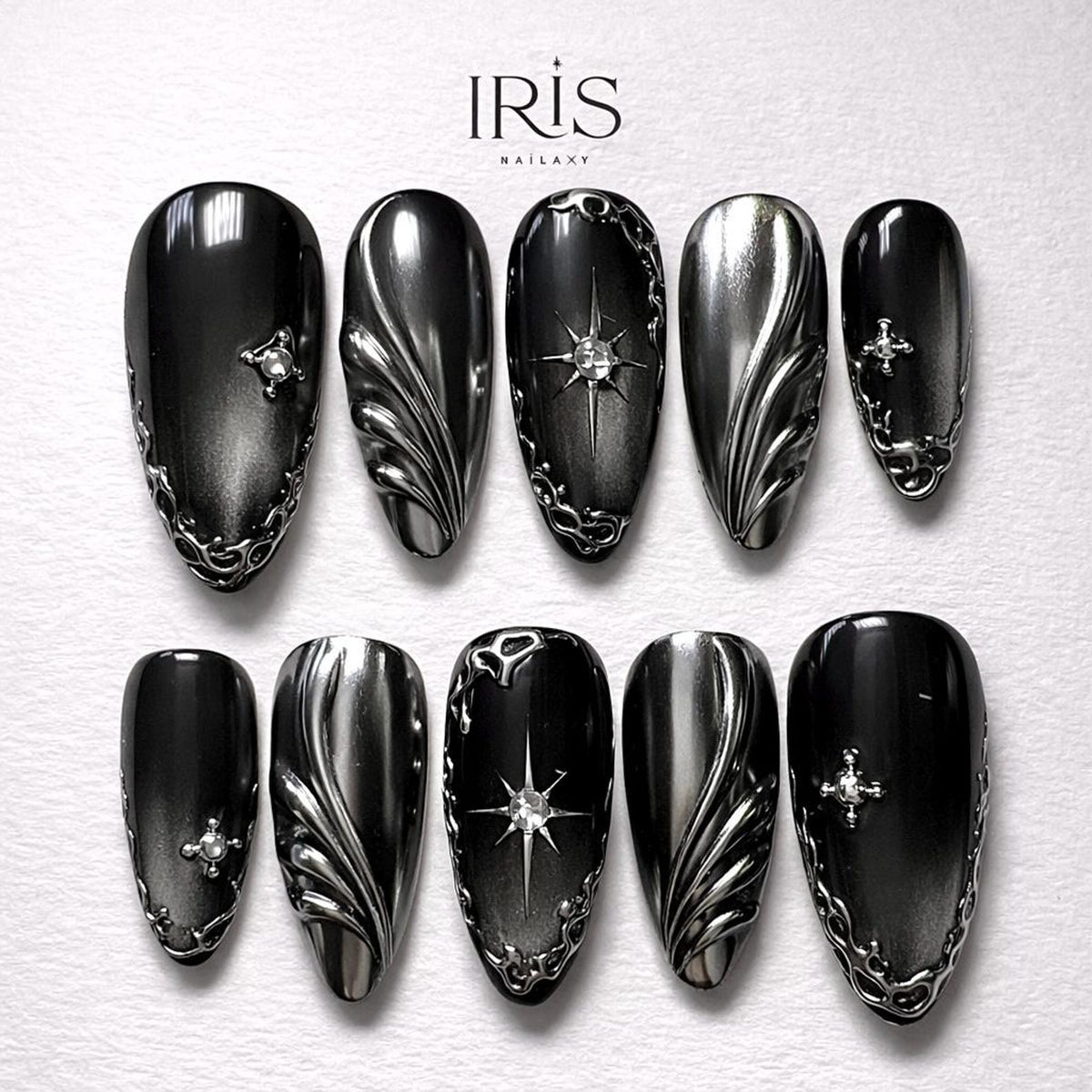 A set of ten black and silver press-on nails. The nails feature various designs, including ornate silver filigree, starburst accents, and small crystal embellishments. The brand name "IRIS" is visible at the top.