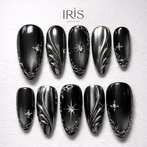 A set of ten black and silver press-on nails. The nails feature various designs, including ornate silver filigree, starburst accents, and small crystal embellishments. The brand name "IRIS" is visible at the top.
