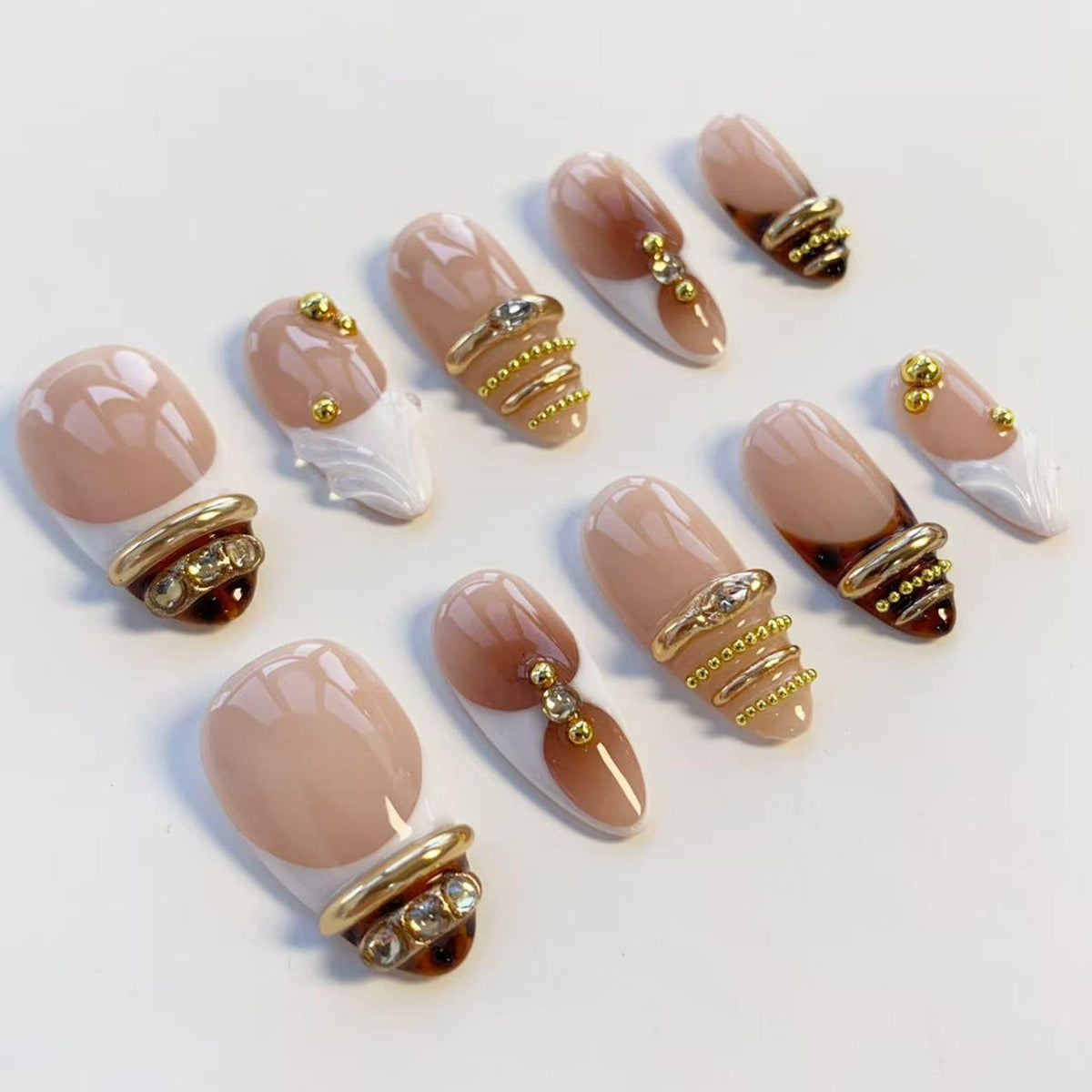 A collection of artificial nails with a nude and brown color scheme. Each nail features gold-colored embellishments, including rings, studs, and decorative elements. Some nails have a white accent.